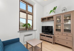 A modern living room with a blue sofa, a TV, a cabinet with glass doors, and a window with a view of buildings. - Kazimierz Riverside Charming Apartment (Kraków)