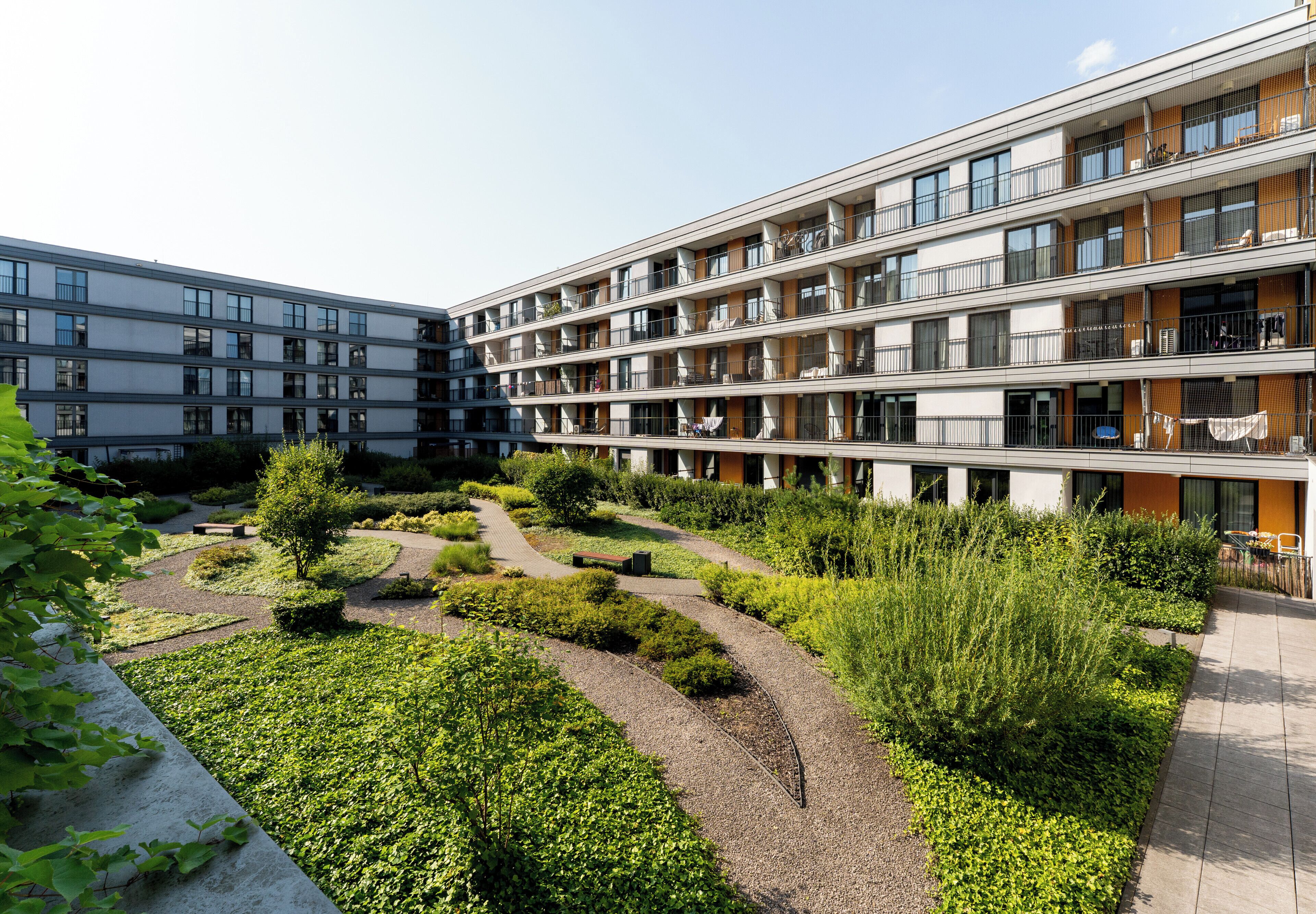 A multi-story residential building with balconies, a landscaped garden area with pathways, and a clear sky.