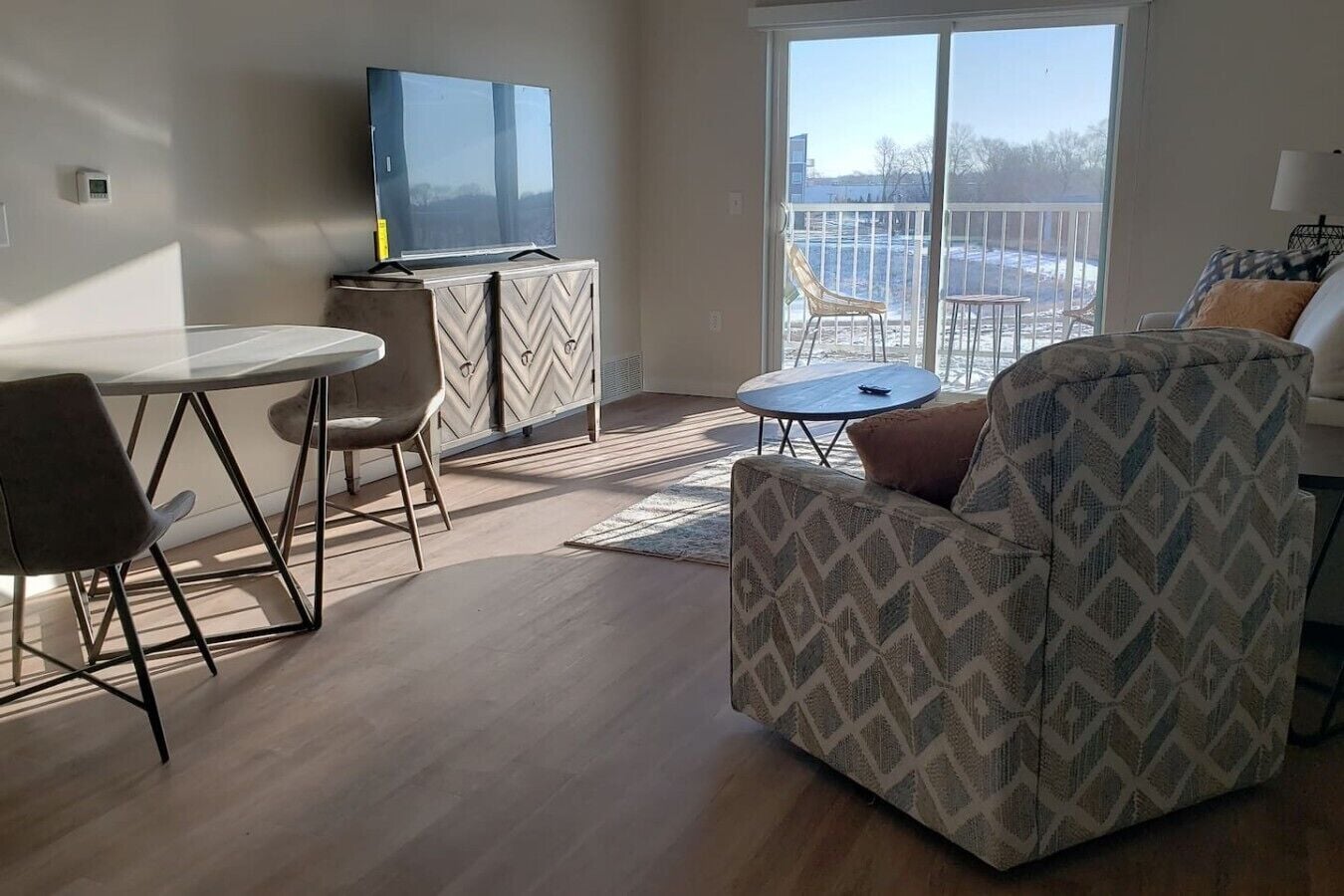 A modern living room with a flat-screen TV, a dining table with chairs, a patterned armchair, and a view of a balcony with outdoor furniture.