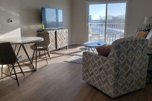 A modern living room with a flat-screen TV, a dining table with chairs, a patterned armchair, and a view of a balcony with outdoor furniture.