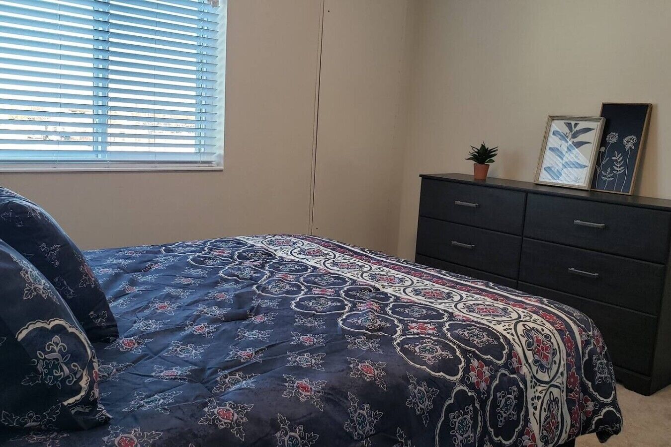 A bedroom with a bed, a dresser, a window with blinds, and a small potted plant.