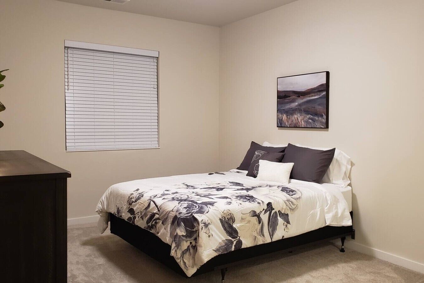 Une chambre à coucher avec un lit, une fenêtre avec des stores, un tableau au mur et un meuble sombre.