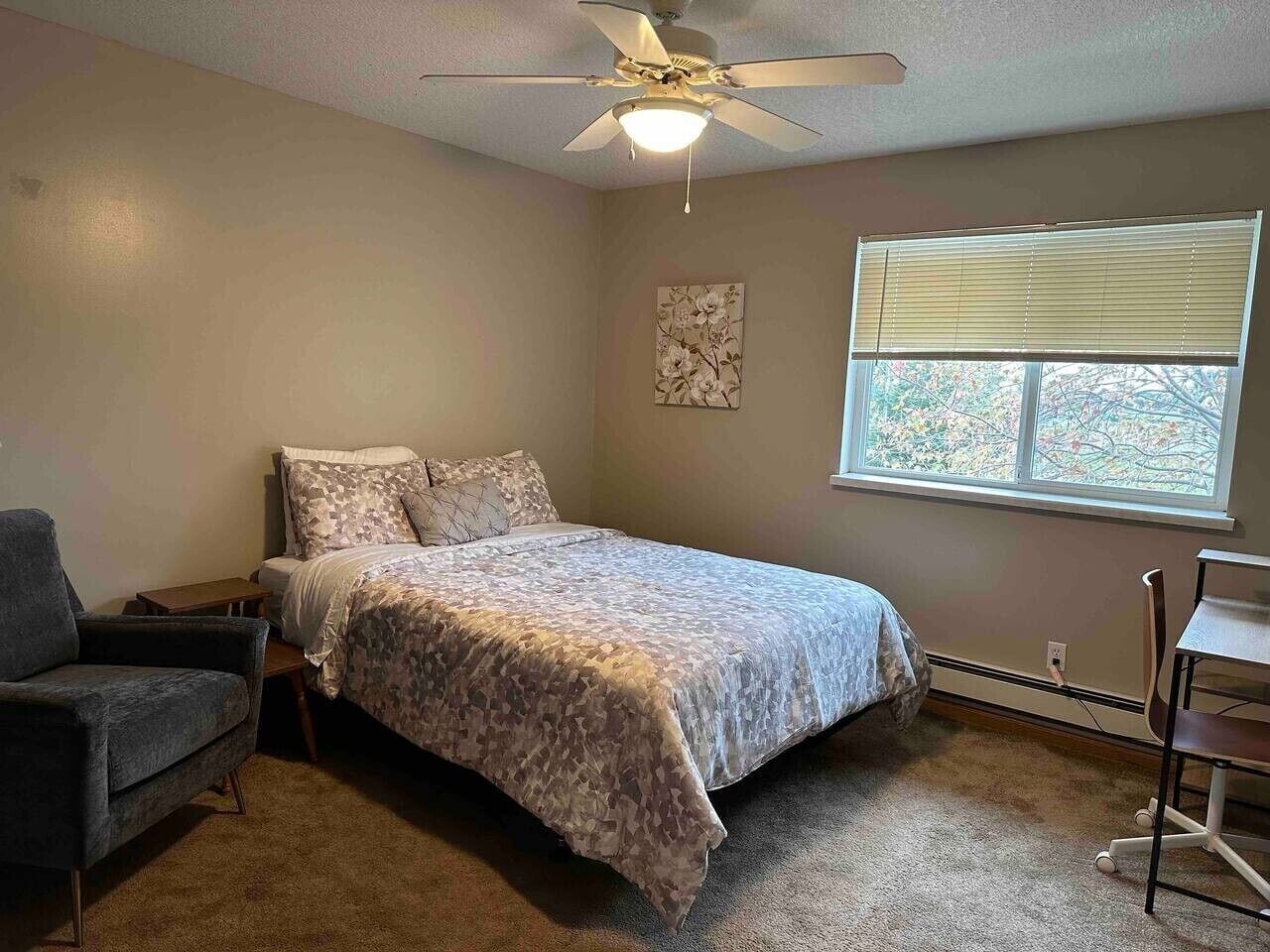 A bedroom with a bed, a chair, a desk, a ceiling fan, a window, and a wall-mounted artwork.
