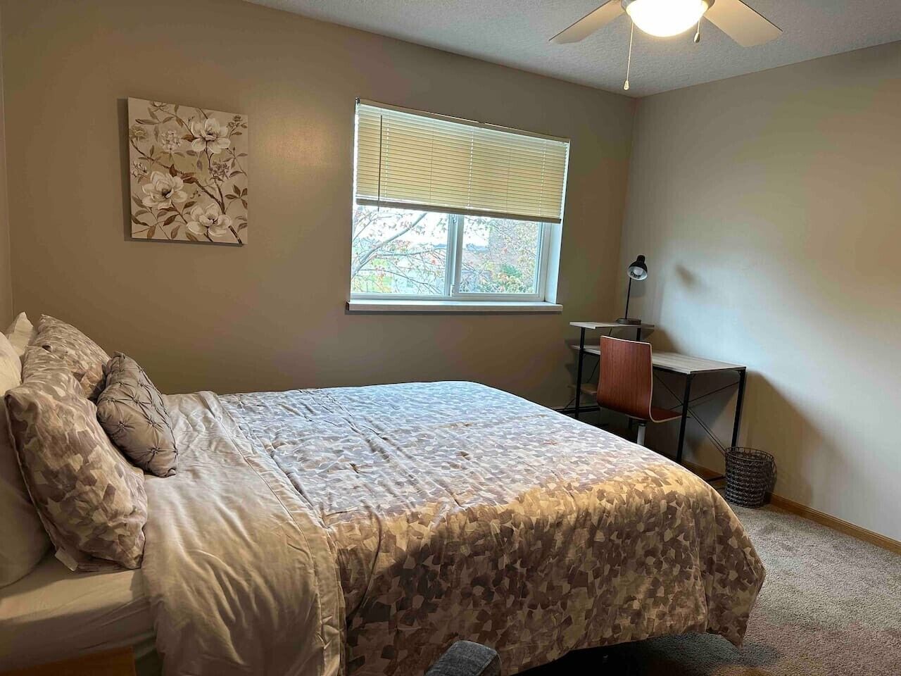 Une chambre à coucher comprenant un lit, un bureau, une chaise, une fenêtre avec des stores et un ventilateur de plafond.