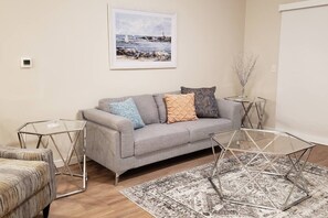 A living room with a grey sofa, two glass tables, a patterned rug, and a framed painting on the wall.