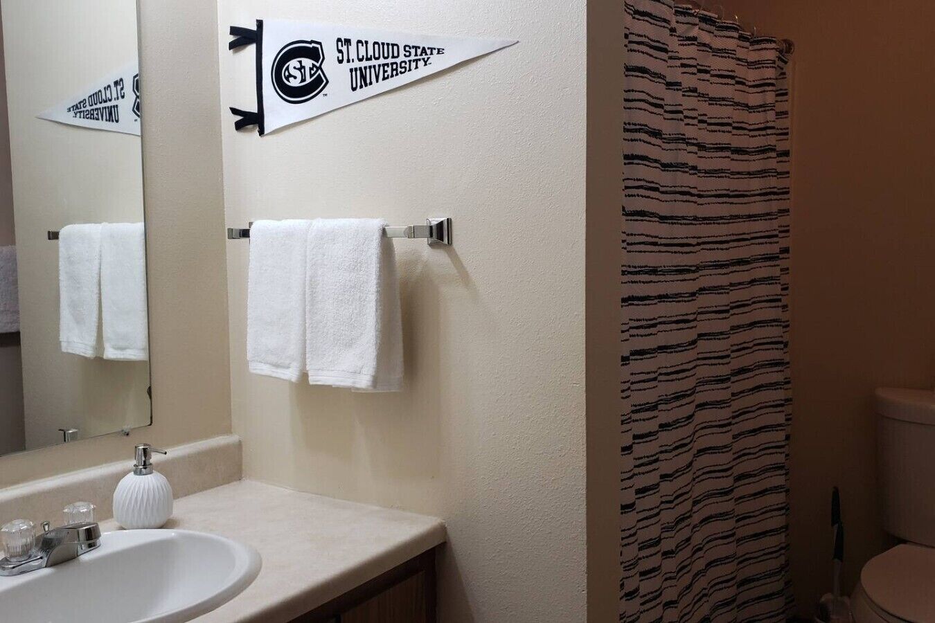 Un baño con lavamanos blanco, dos toallas blancas, cortina de ducha a rayas y un cartel de la Universidad Estatal de St. Cloud.