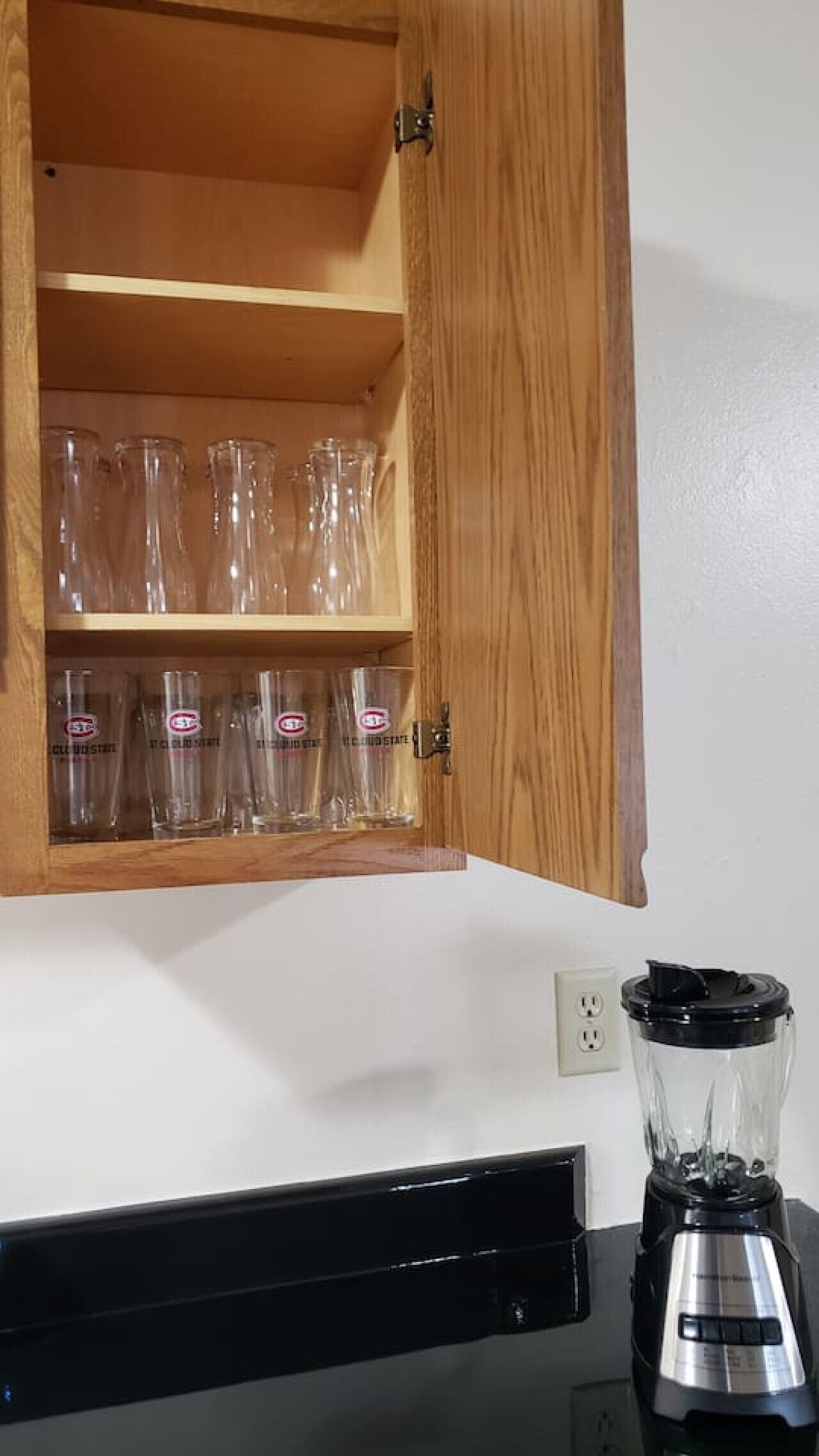 A kitchen with a wooden cabinet containing clear drinking glasses and a black blender on the counter.
