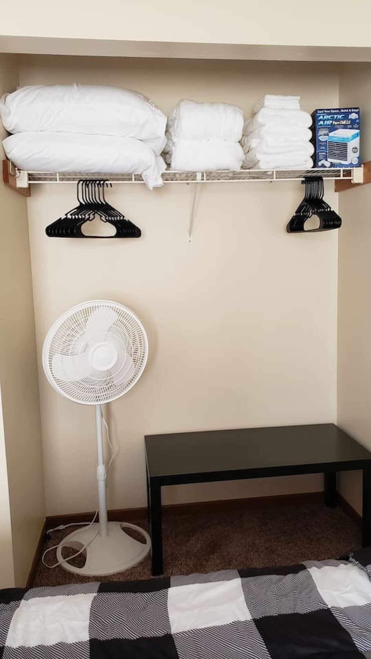 Una habitación con una cama, un ventilador de pie blanco, un banco negro y un estante con toallas dobladas y una caja.