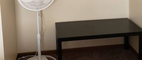 A room with a bed, a white standing fan, a black bench, and a shelf with folded towels and a box.