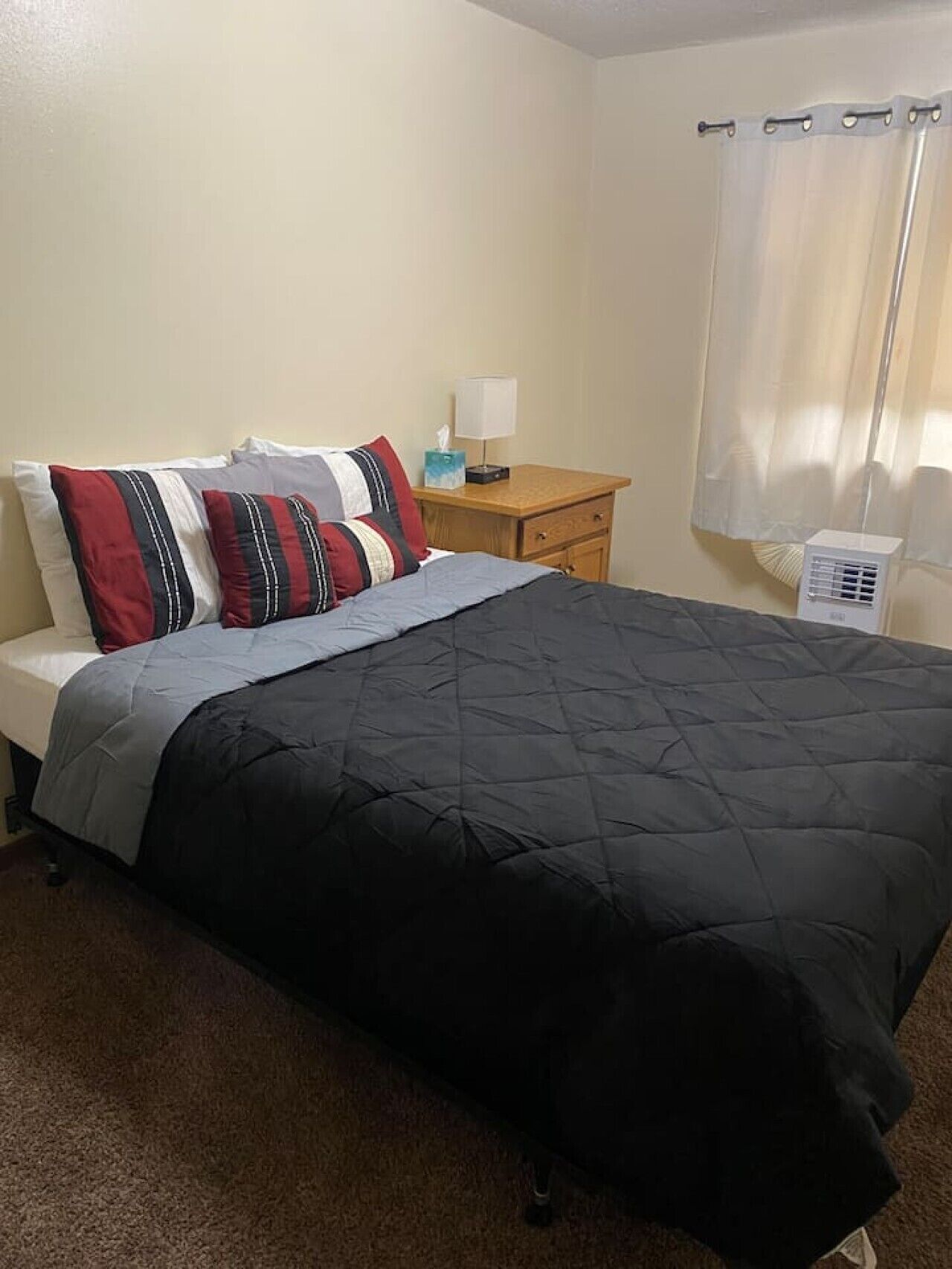 A single bed with a black quilted cover, a wooden nightstand with a lamp, and a white air purifier.