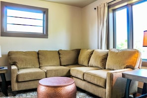 A corner living room with a beige sectional sofa, a round copper coffee table, and a small side table. - Historic 4 BR Copper Mine House/ Sauna /Near Trails & Downtown Calumet (Laurium)