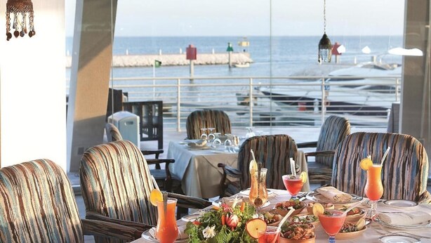 Une table à manger dressée pour un repas, avec une vue sur la mer et des bateaux en arrière-plan.