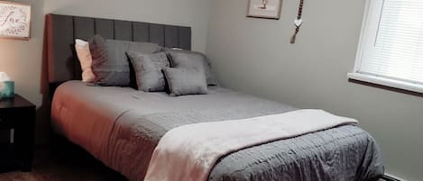 A bedroom with a bed, pillows, a bedside table, a framed picture, and a window with blinds.