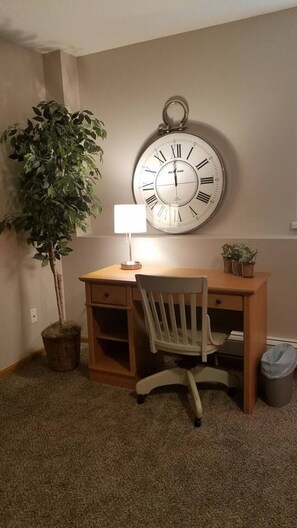 A room with a wooden desk, a chair, a wall-mounted clock, a potted plant, and a trash bin.