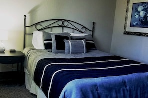 Una cama bien hecha con una colcha azul y blanca a rayas, un cabecero de metal y una mesita de noche con una lámpara.