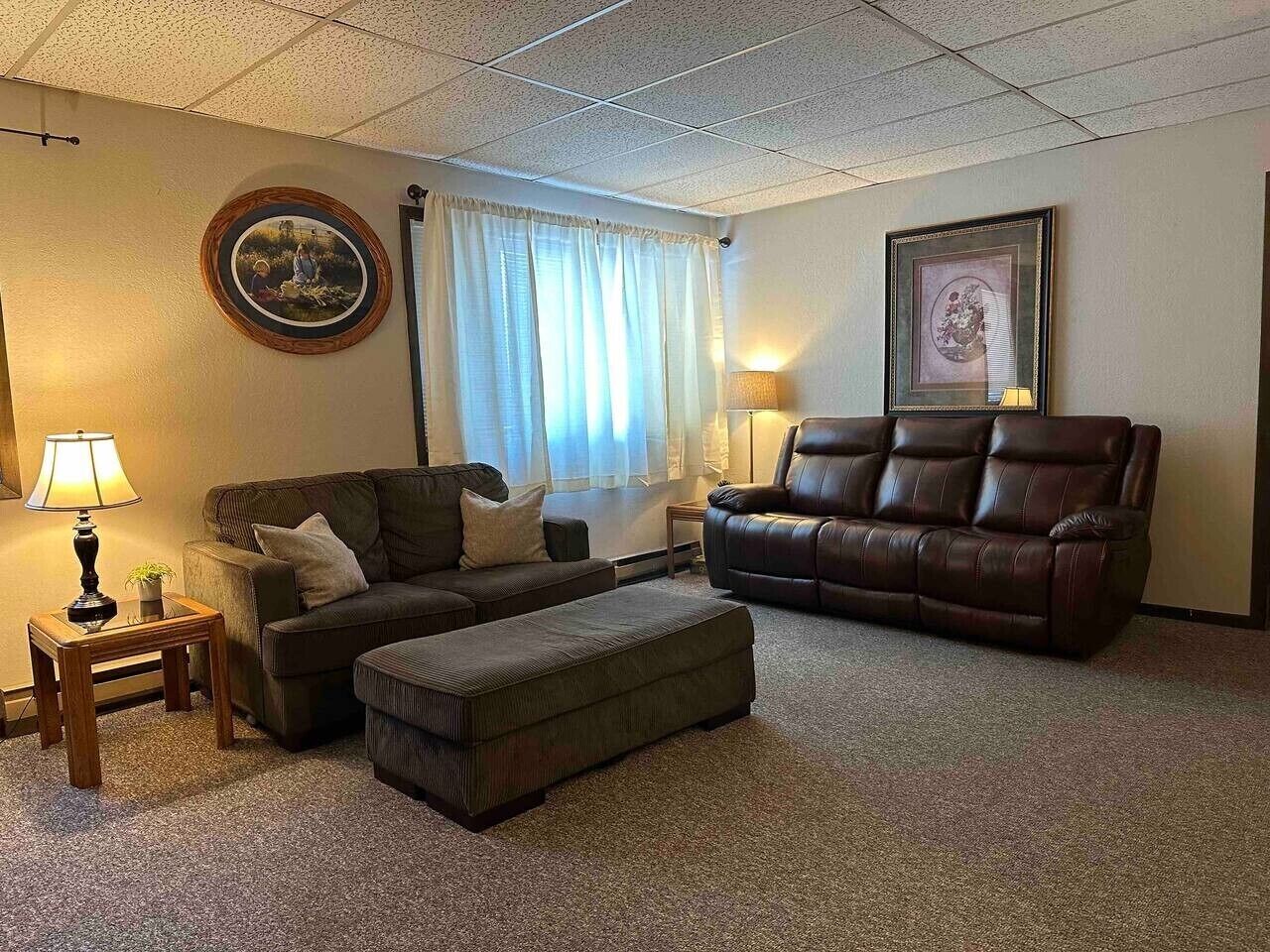 A living room with a brown leather sofa, a matching ottoman, a wooden side table with a lamp, and two framed pictures on the walls.