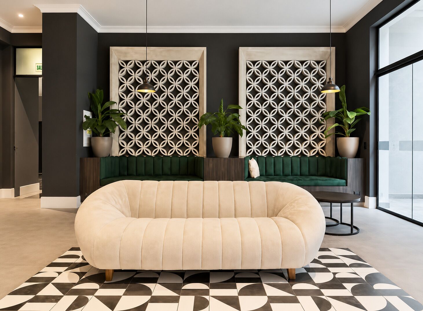A modern living room with a beige sofa, a dark green sofa, and a patterned wall divider. There are two potted plants and a small table with a lamp.