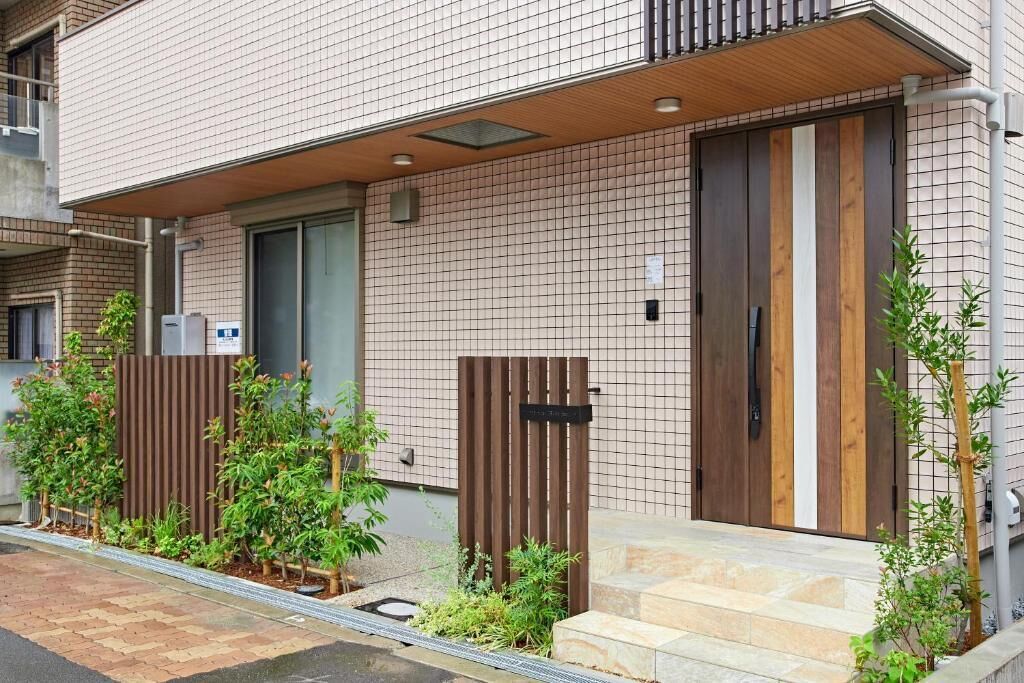 A modern building with a tiled facade, a wooden door, and a small garden area with plants.