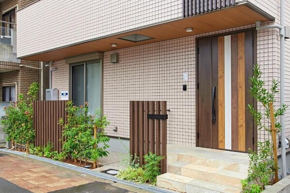 A modern building with a tiled facade, a wooden door, and a small garden area with plants.
