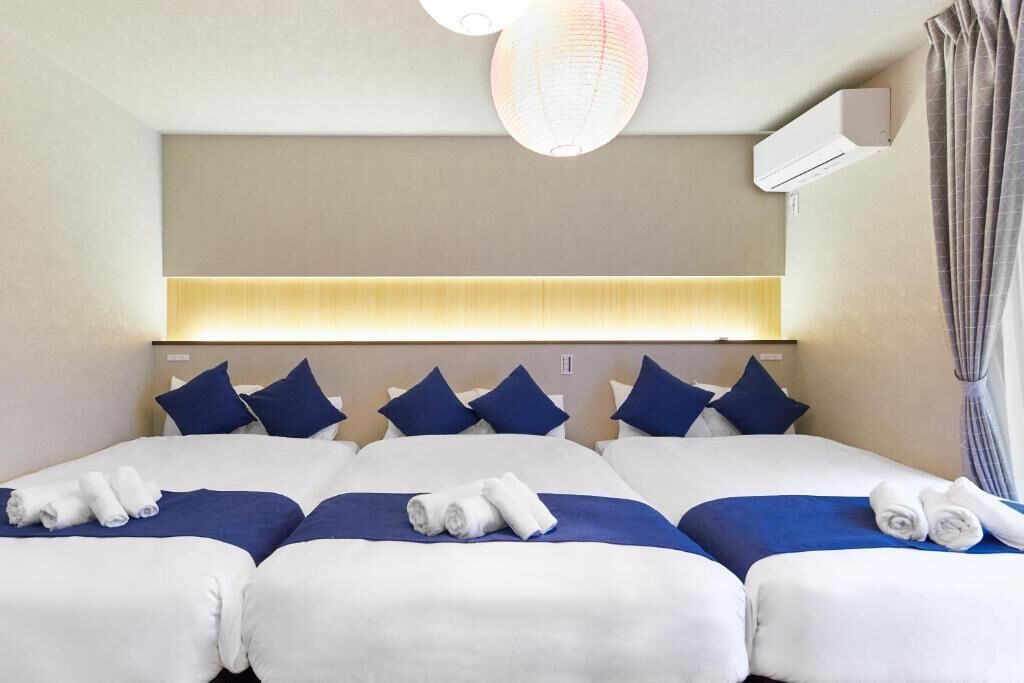 A hotel room with a large bed, multiple pillows, a ceiling light fixture, an air conditioning unit, and a window with curtains.