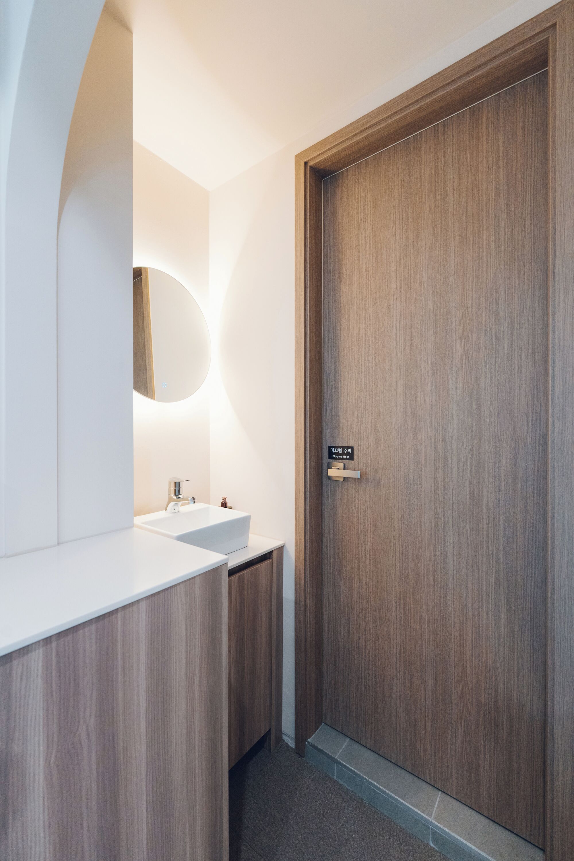 Un baño moderno con puerta de madera, espejo con luz circular y lavamanos blanco.