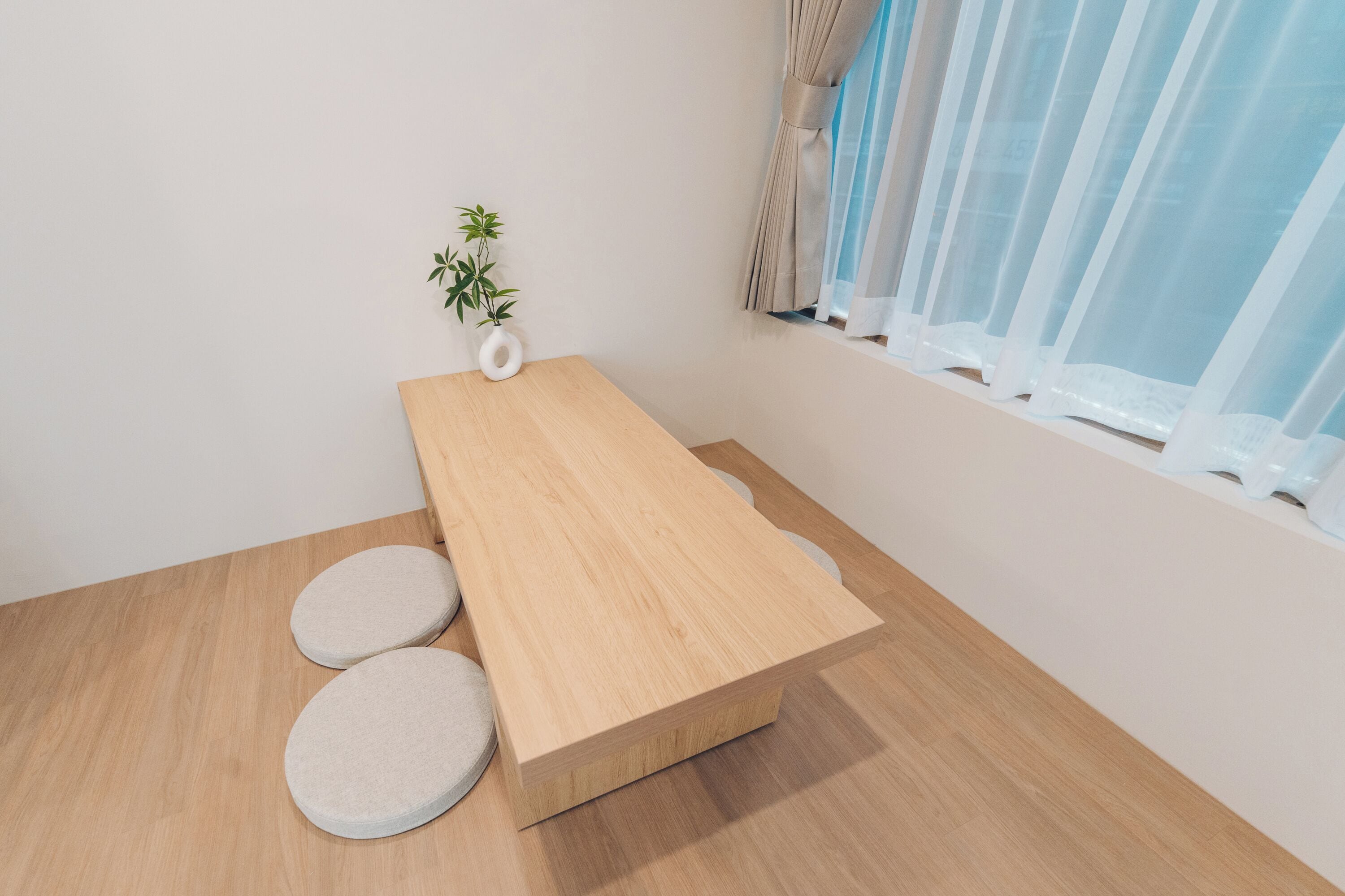 Una habitación minimalista con una mesa de madera, dos taburetes redondos, una pequeña planta en maceta y una ventana con cortinas traslúcidas.