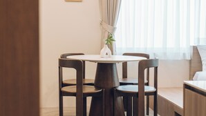 Une chambre d’hôtel avec une table ronde, quatre chaises, une plante, un tableau encadré au mur et une fenêtre avec des rideaux.