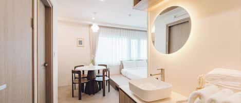 Una habitación de hotel con un espejo redondo, una cama, una mesita de noche, una mesa redonda con sillas y un ventanal con cortinas.