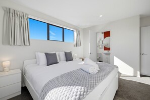 A modern bedroom with a large bed, a bedside table, a nightstand, a window with curtains, and a bathroom with a sink and mirror. - Entire home in Christchurch, New Zealand (Christchurch)