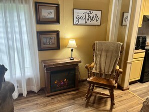 A cozy living room with a fireplace, a wooden chair with a towel, framed pictures on the wall, and a 'Gather' sign. - Camp / 1097 Elm run (St. Martin Parish)
