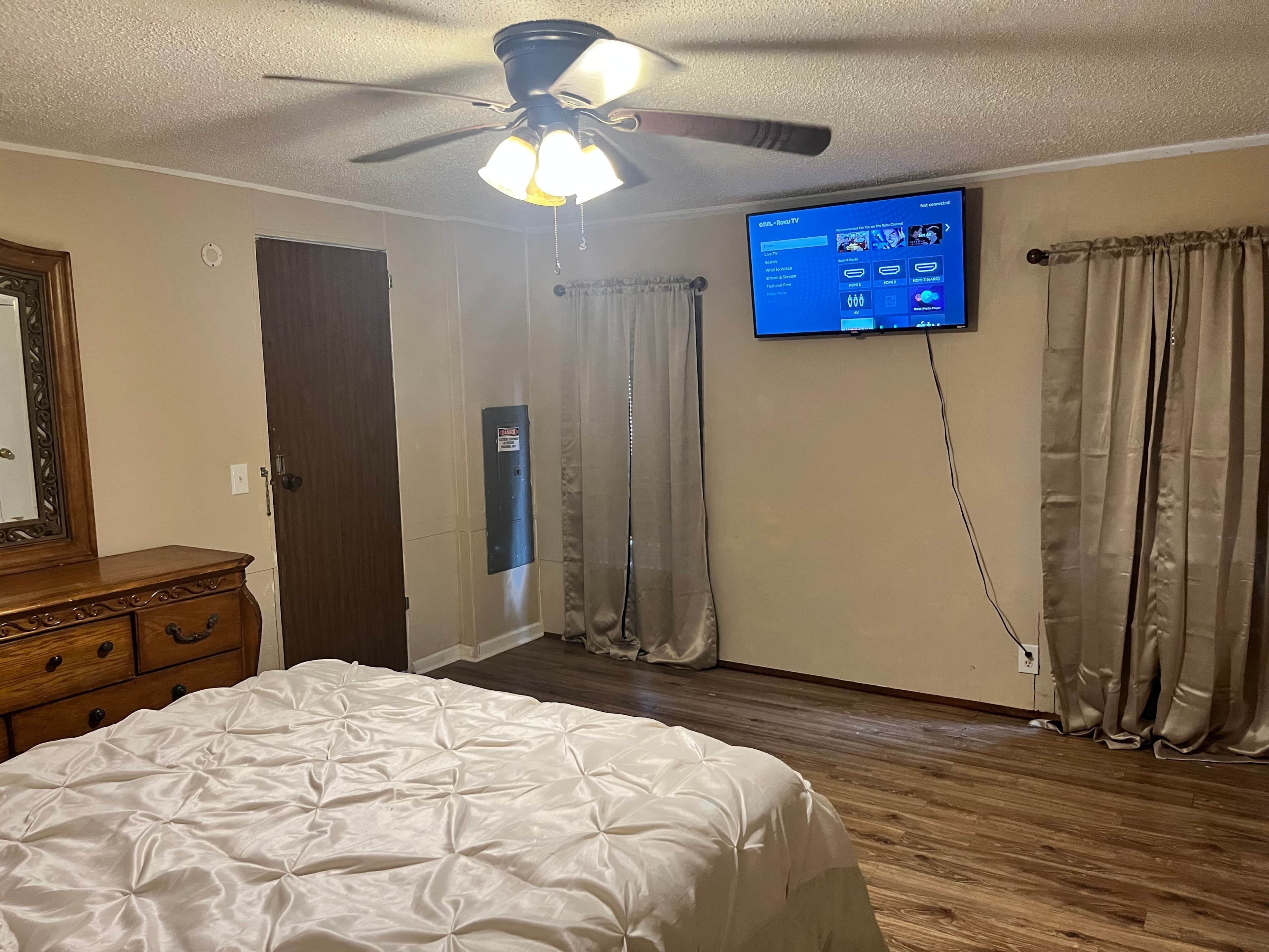 A bedroom with a bed, a ceiling fan, a television mounted on the wall, and a wooden dresser.