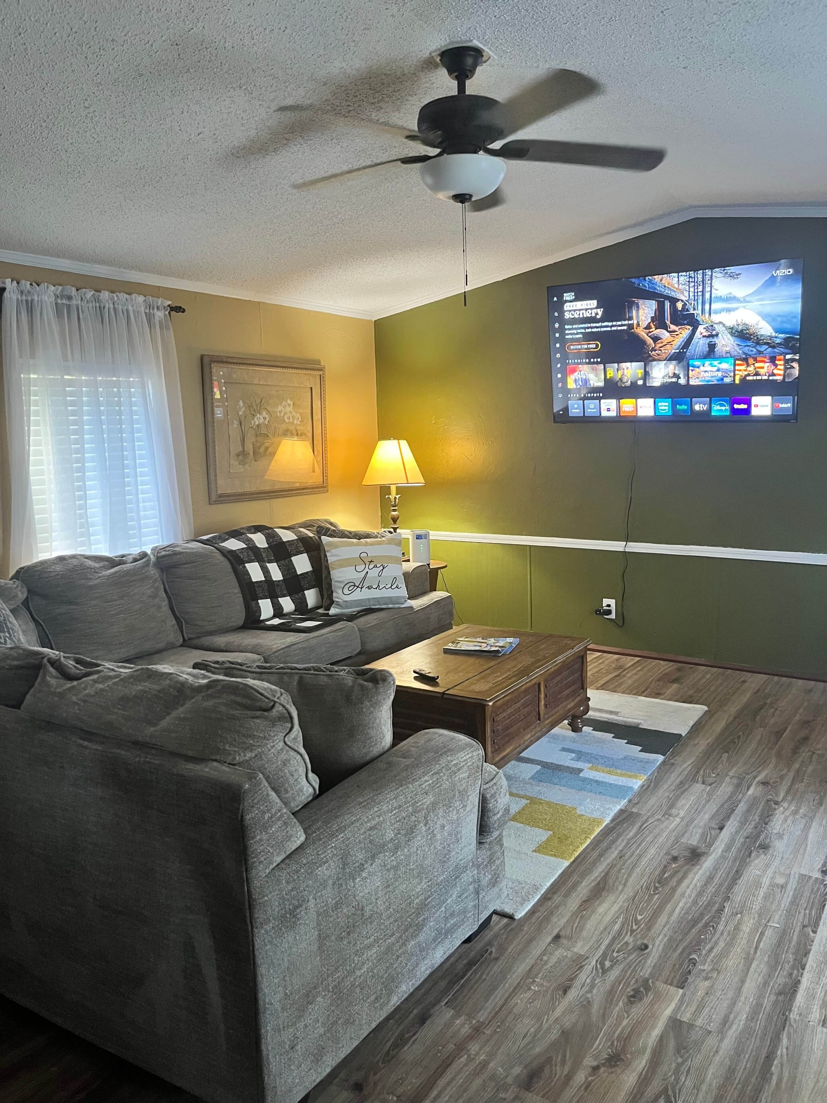 A living room with a sofa, a coffee table, a ceiling fan, a TV mounted on the wall, and a lamp.