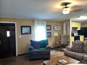 A living room with a sofa, armchair, and a coffee table. There is a door, a window with curtains, and a ceiling fan. - Camp / 1097 Elm run (St. Martin Parish)
