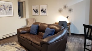 A living room with a brown leather sofa, two blue cushions, a wooden coffee table, a lamp, and wall art.