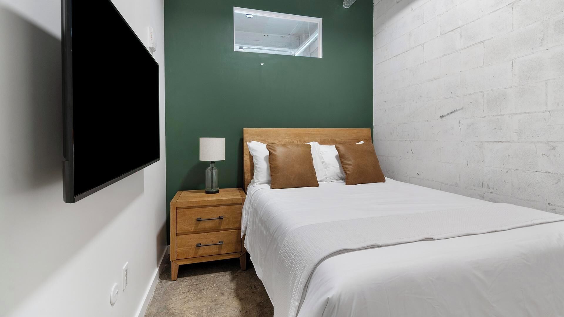 Een slaapkamer met een bed, nachtkastje, lamp, raam en een aan de muur bevestigde televisie.