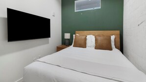 Hotellihuone, jossa on sänky, seinälle asennettu televisio, yöpöytä lampun kanssa ja ikkuna, jossa on ilmastointilaite.