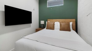 A hotel room with a bed, a TV mounted on the wall, a nightstand with a lamp, and a window with an air conditioning unit. - Landing | Spectacular 1BD, Rooftop Lounge, Gym (Jacksonville)