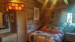 A wooden cabin bedroom with a bed, a dresser, and a window with lace curtains.