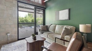 A living room with a sofa, a coffee table, and a lamp. There is a window with a view of a parking area and greenery outside. - Landing | Modern 1BD, Gym, Rooftop Lounge (Jacksonville)