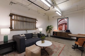 Ein modernes Wohnzimmer mit einer Couch, einem runden Couchtisch, einem Beistelltisch, einem kleinen Hocker, einer Topfpflanze, einem Fernseher und einem Esstisch mit Stühlen.