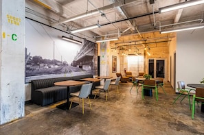 Een modern café met zitplaatsen, openliggende leidingen in het plafond en een muurschildering.