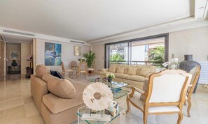 Ein geräumiges Wohnzimmer mit einem beigen Sofa, einem Glastisch und einem Holzstuhl. Es gibt einen Essbereich mit einem Glastisch und Stühlen, sowie ein großes Fenster mit Blick ins Freie.