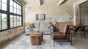 A room with a large window, a sofa, a coffee table, a chair, and a small table with a lamp. - Landing | Beautiful 1BD, Gym, Rooftop Lounge (Jacksonville)