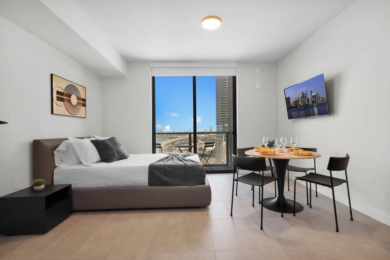 Un dormitorio moderno con cama, una mesa de comedor con sillas, vista a la ciudad y obras de arte en las paredes.