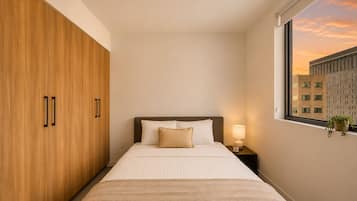 Une chambre d’hôtel avec un lit, une table de chevet, une fenêtre donnant sur des immeubles et des portes de placard en bois.