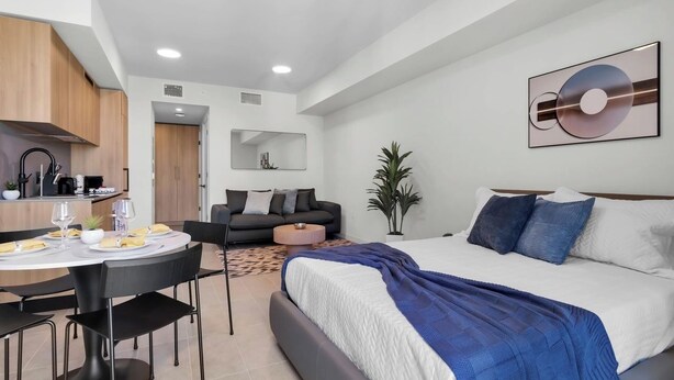 A modern hotel room with a bed, a dining area with a table and chairs, a sofa, a small round table, and a potted plant. - Miami Luxe Suites (Miami)