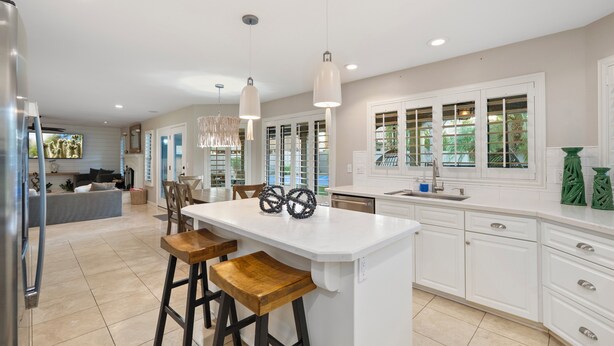 A modern kitchen with white cabinetry, a central island with bar stools, pendant lighting, and a view of the outdoors through large windows. - Luxury 5BR Retreat, Heated Pool, Spa, Sport Court, Putting Green & Game Rooms (Scottsdale)