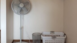A laundry area with a fan, a shelf with folded towels, a basket, and a red cloth.