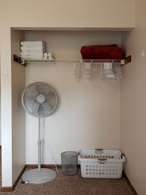A laundry area with a fan, a shelf with folded towels, a basket, and a red cloth.