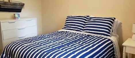 A bed with a striped bedspread, a white bedside table with a lamp, and a white dresser with drawers.